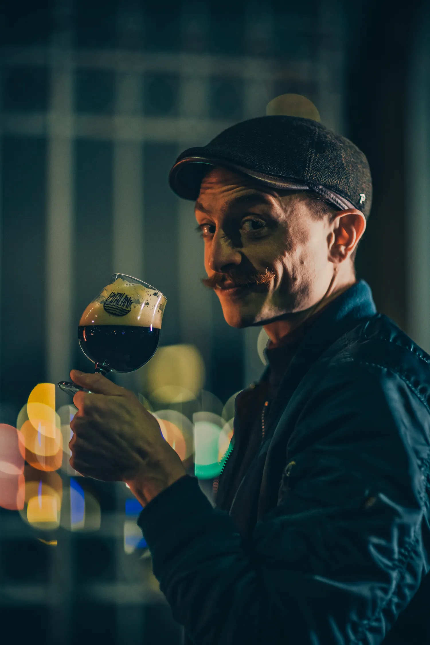 Man in a flat cap smiling over a glass of dark beer in a pub at night, Athletic Brewing Company