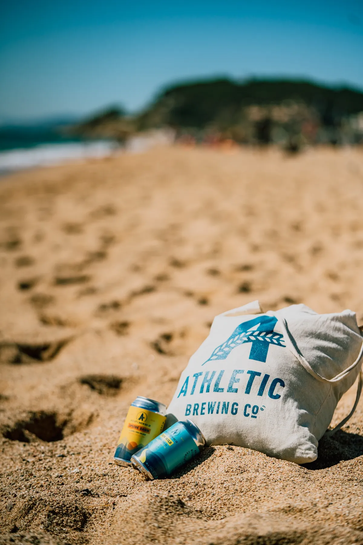 Two cans and a white Athletic Brewing tote bag lying on a sandy beach, the sea behind, brand shoot