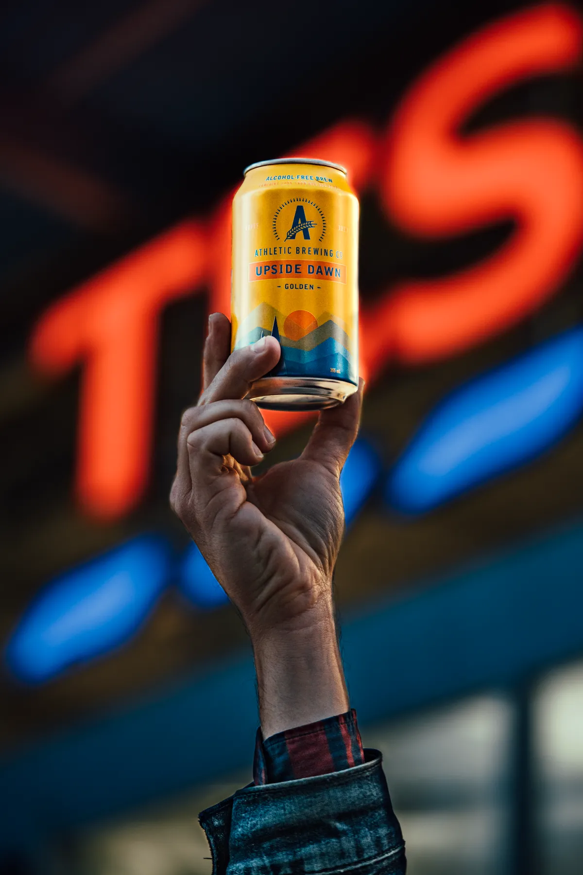 Hand raising a yellow Upside Dawn can against a red and blue neon sign, Athletic Brewing Company