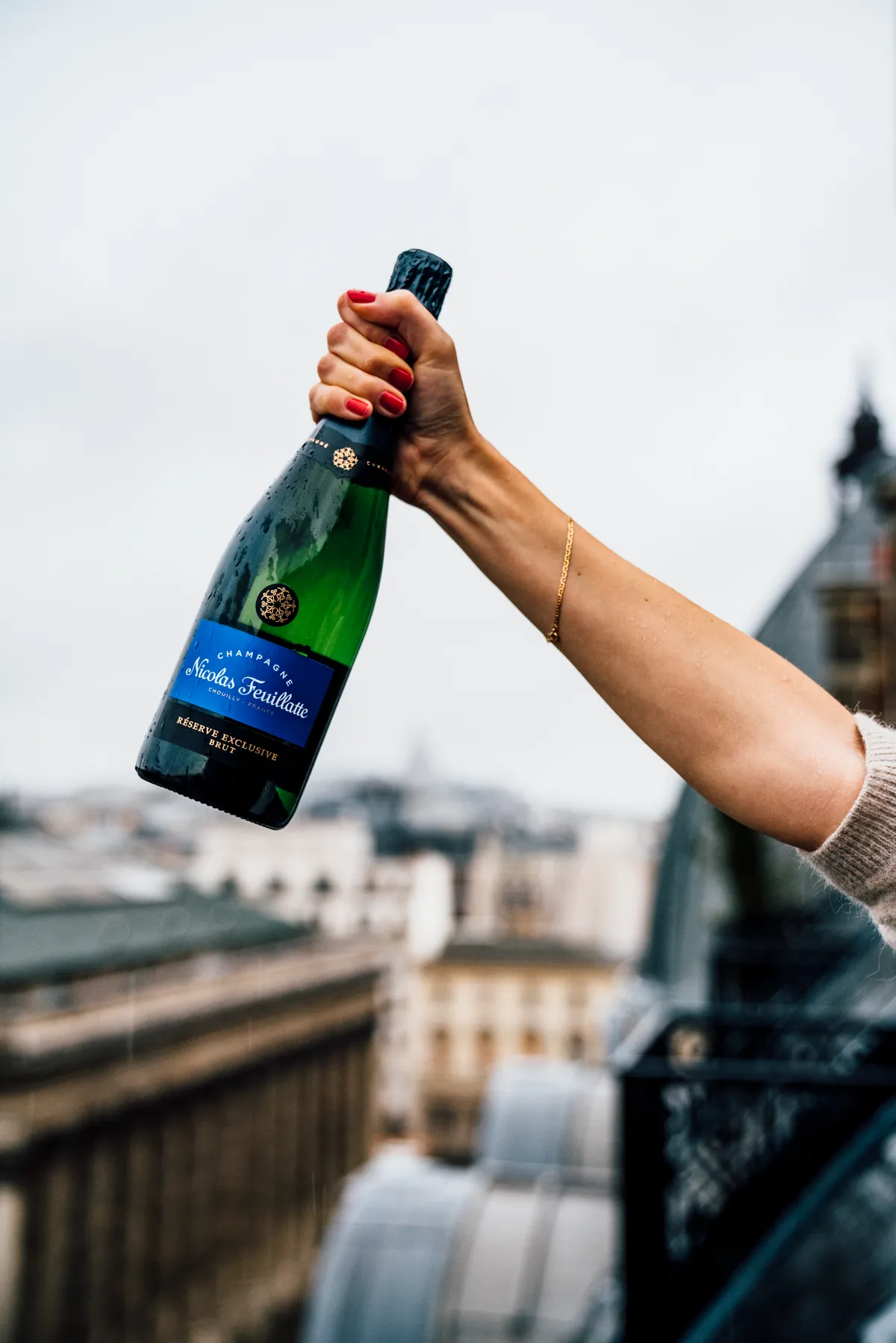 Hand holding a blue-label bottle of Nicolas Feuillatte champagne above the rooftops of Paris