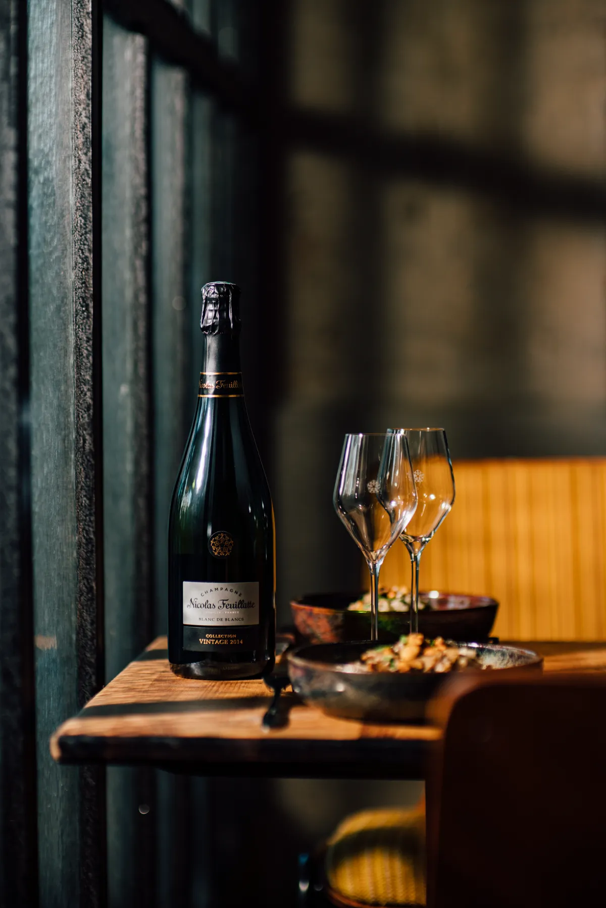 Bottle and two glasses on a small table in a dim restaurant corner, Nicolas Feuillatte champagne