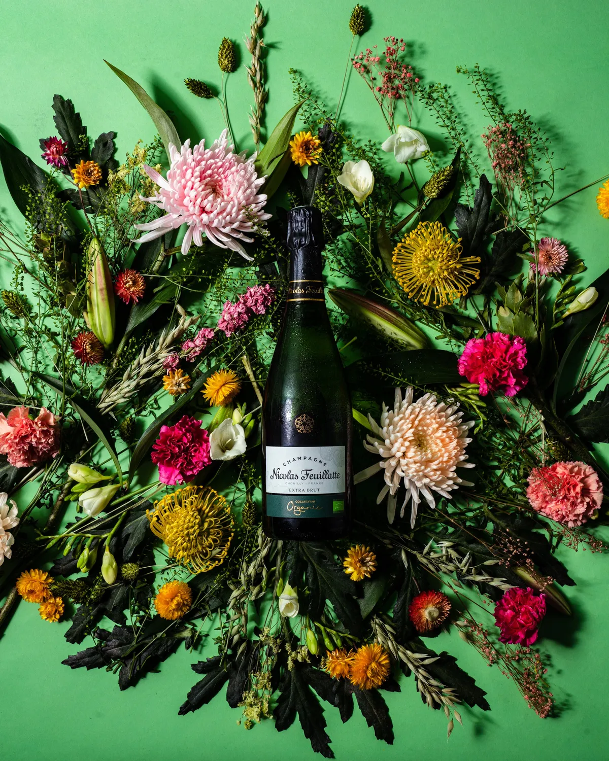 Bottle of Nicolas Feuillatte champagne lying in a wreath of colourful flowers on a green background