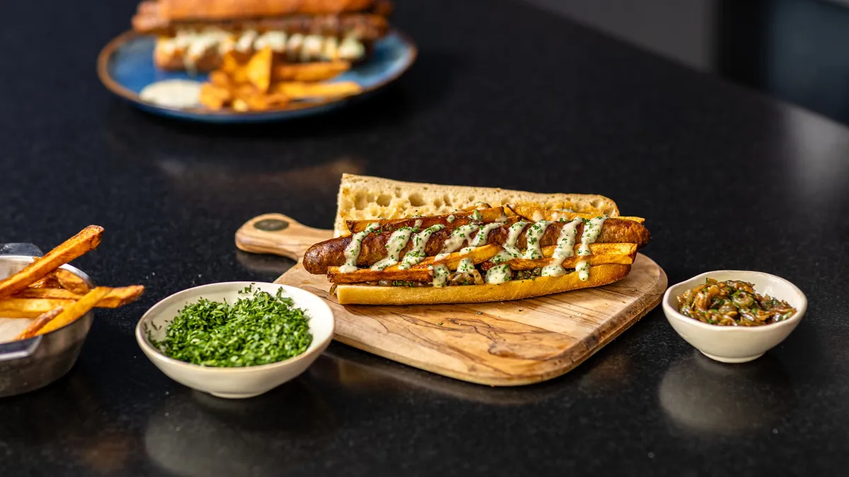 Merguez sandwich with herb sauce on a wooden board, small bowls around it, food shot for Graille sur le rail