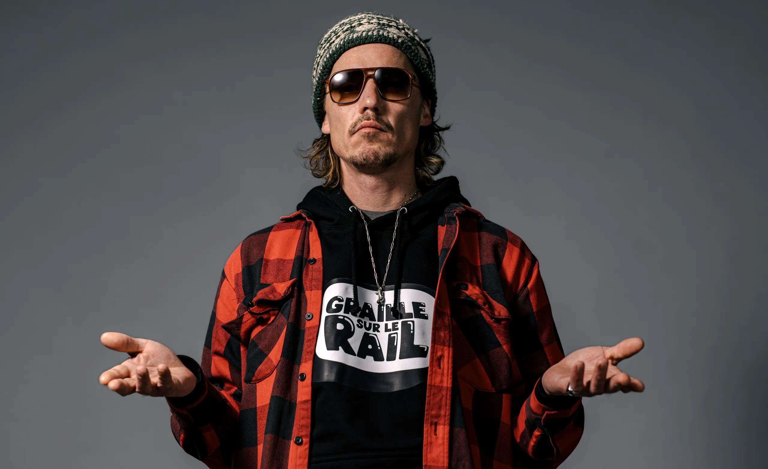 Scotchman in a beanie, sunglasses and plaid shirt, shrugging, the show's logo on his T-shirt, Graille sur le rail