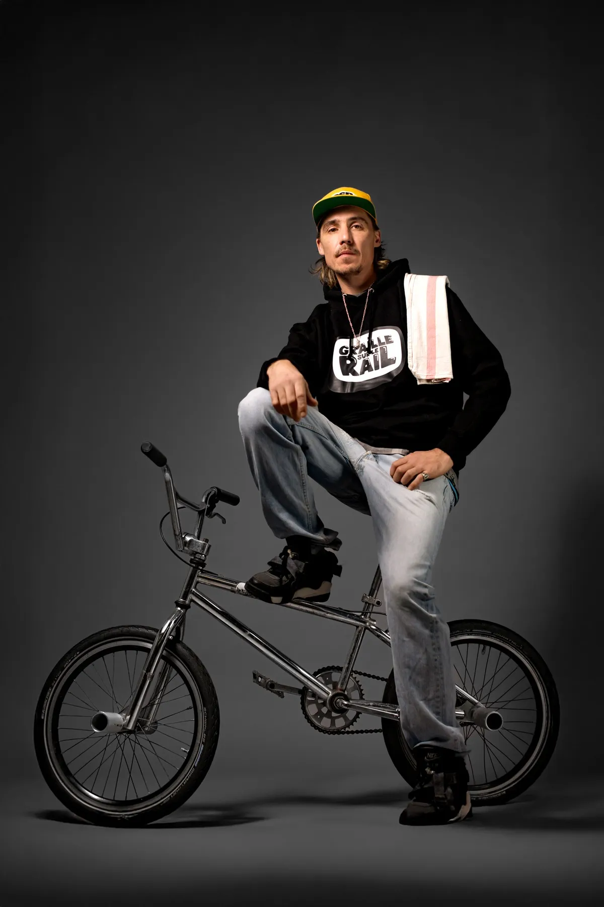 Scotchman sitting on a BMX in the show's hoodie, a paper bag in hand, studio portrait for Graille sur le rail