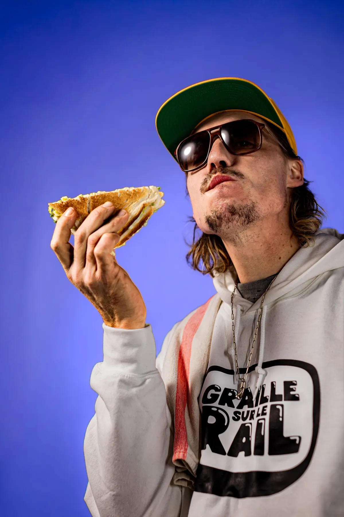 Scotchman in the show's hoodie about to bite into a sandwich against a blue background, Graille sur le rail
