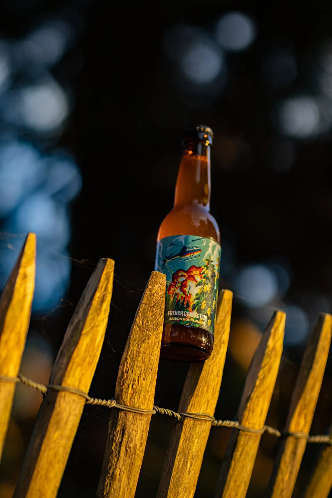 Bottle of French Connection standing on a wooden fence at night, lights blurred behind, Hoppy Road brewery