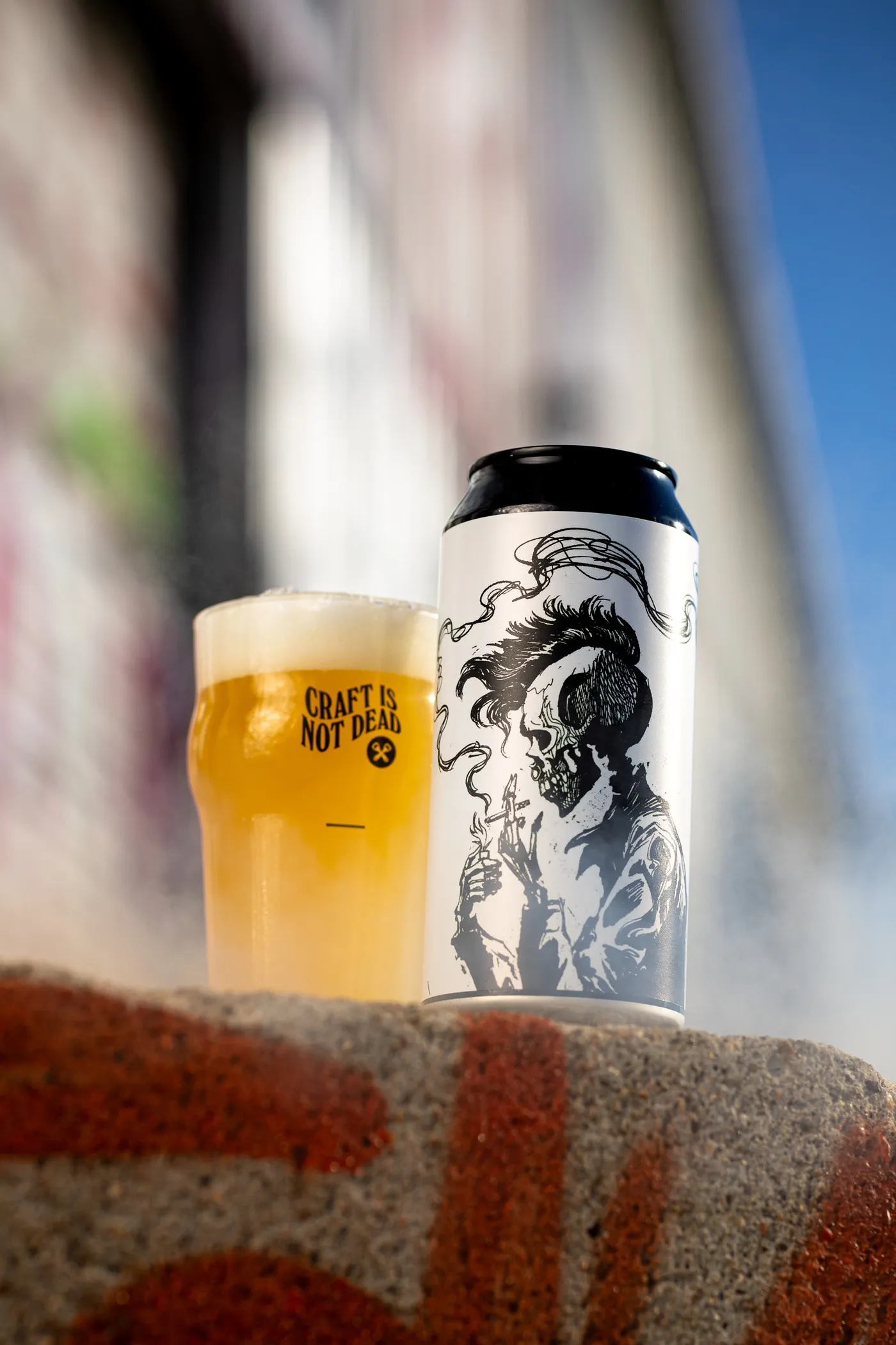 Pint and black and white illustrated can on a graffiti-covered wall, blue sky behind, Hoppy Road brewery