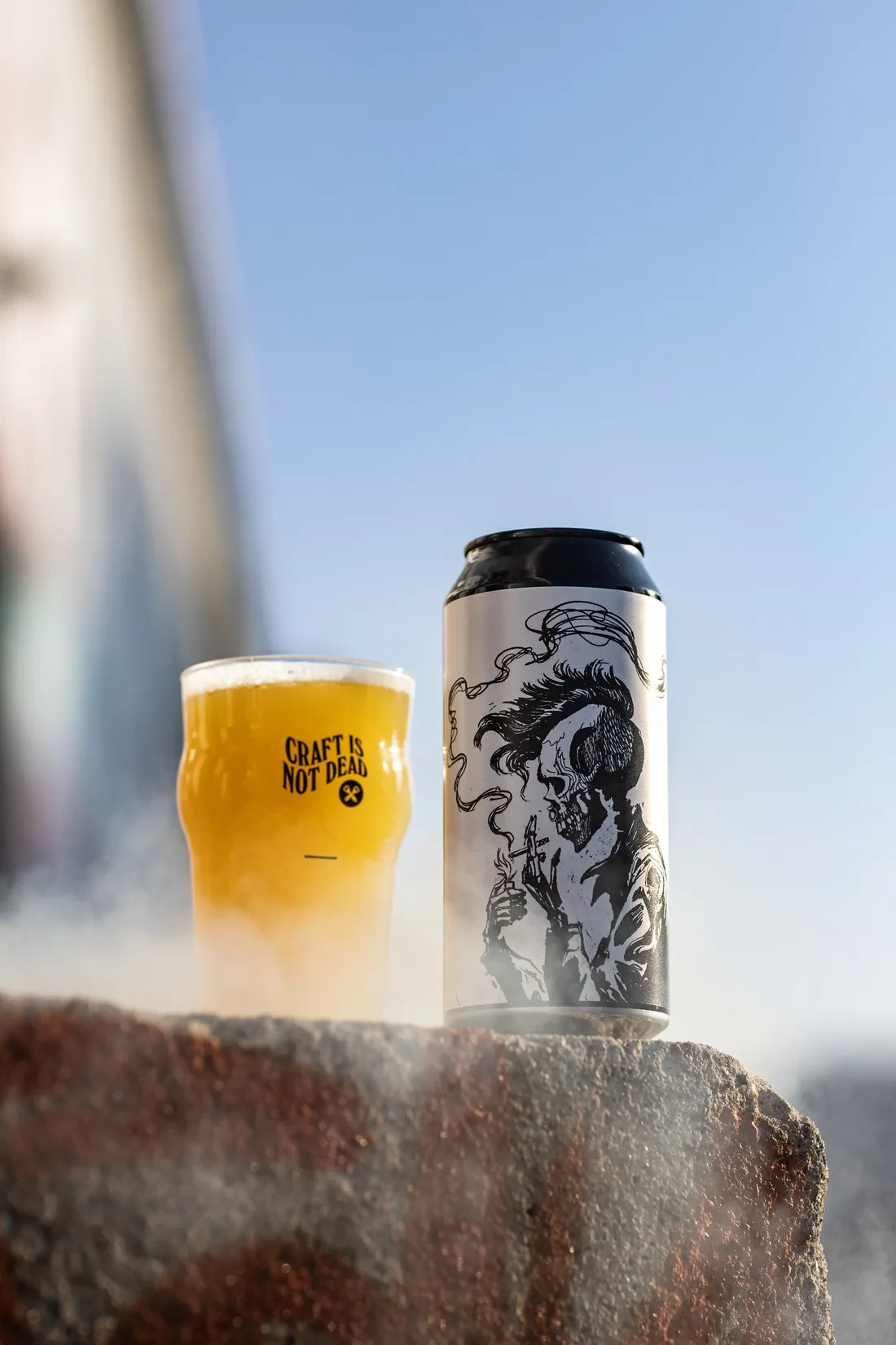 Black and white illustrated can and a pint on a wall ledge under a clear blue sky, Hoppy Road brewery