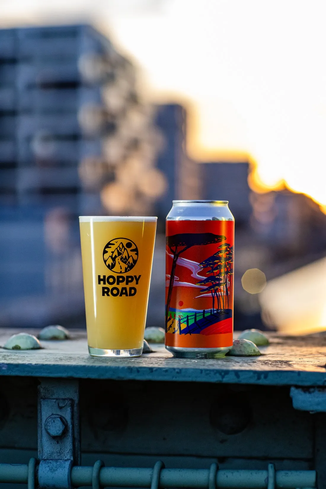 Pint and red illustrated can on a metal railing, buildings blurred in the evening light, Hoppy Road brewery