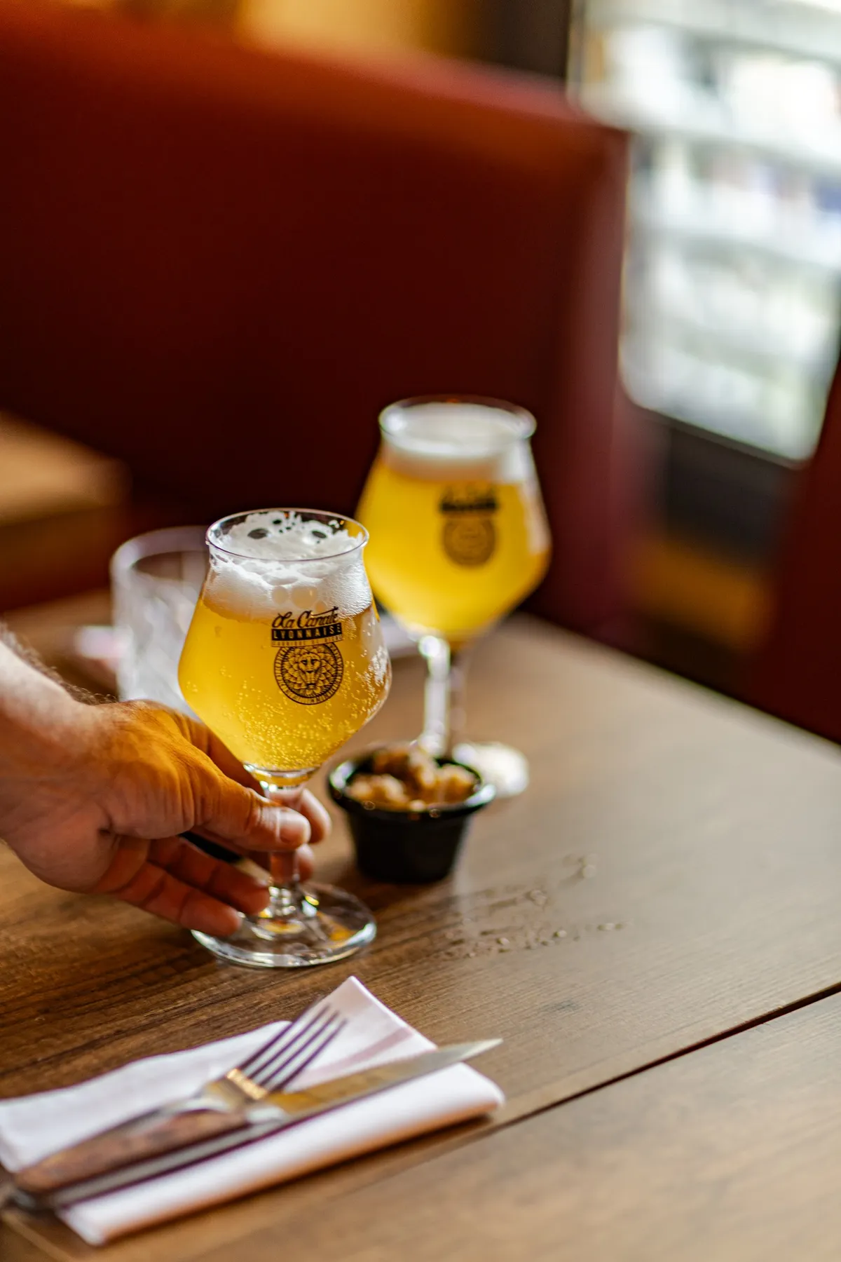 Hand setting a glass of beer on a bistro table next to a second glass and a small bowl, La Canute Lyonnaise