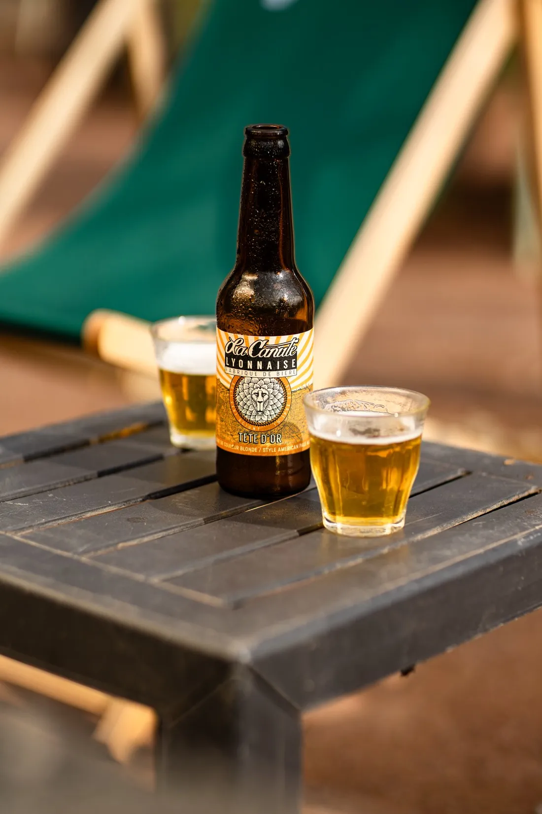 Bottle and two glasses of beer on a small table next to a green deckchair, La Canute Lyonnaise