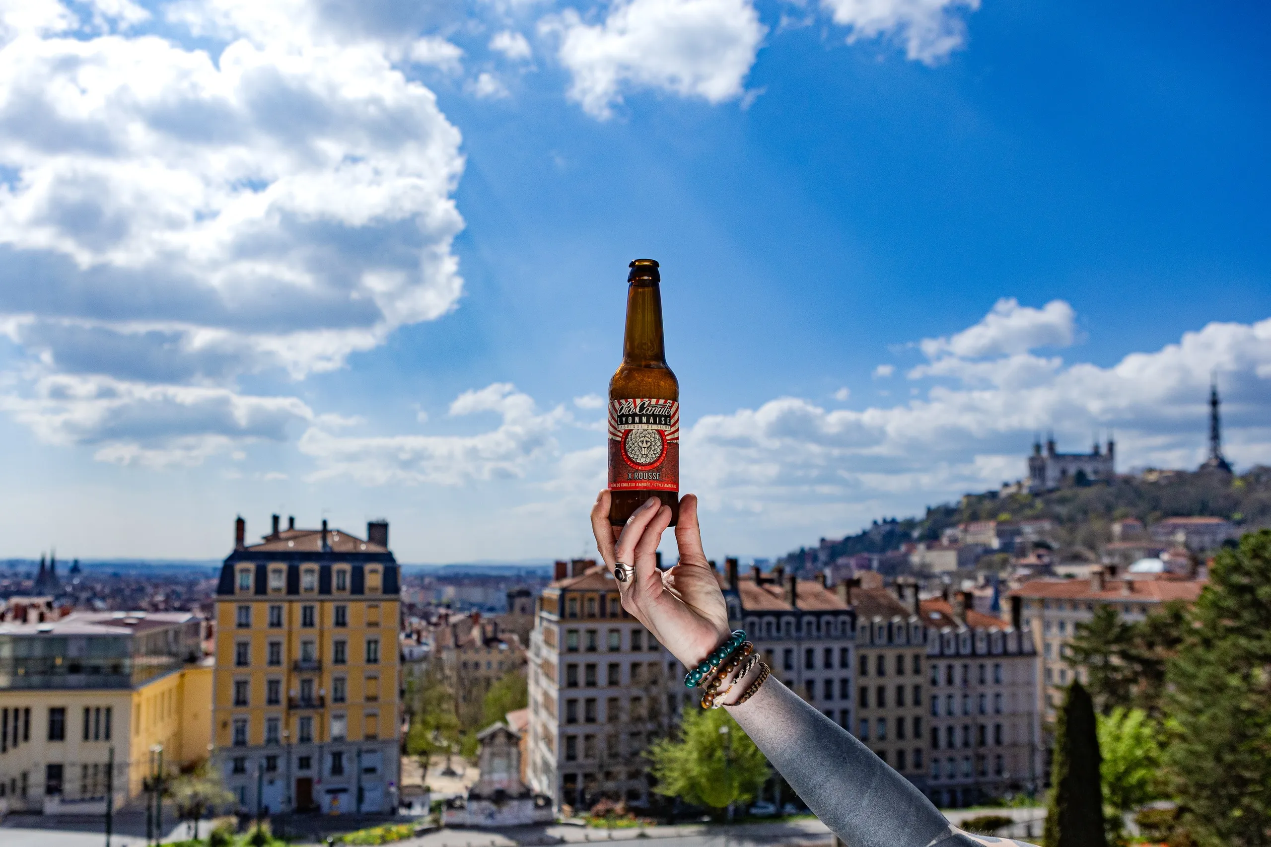 Hand raising a bottle above the rooftops of Lyon, the Fourvière basilica on the hill, La Canute Lyonnaise