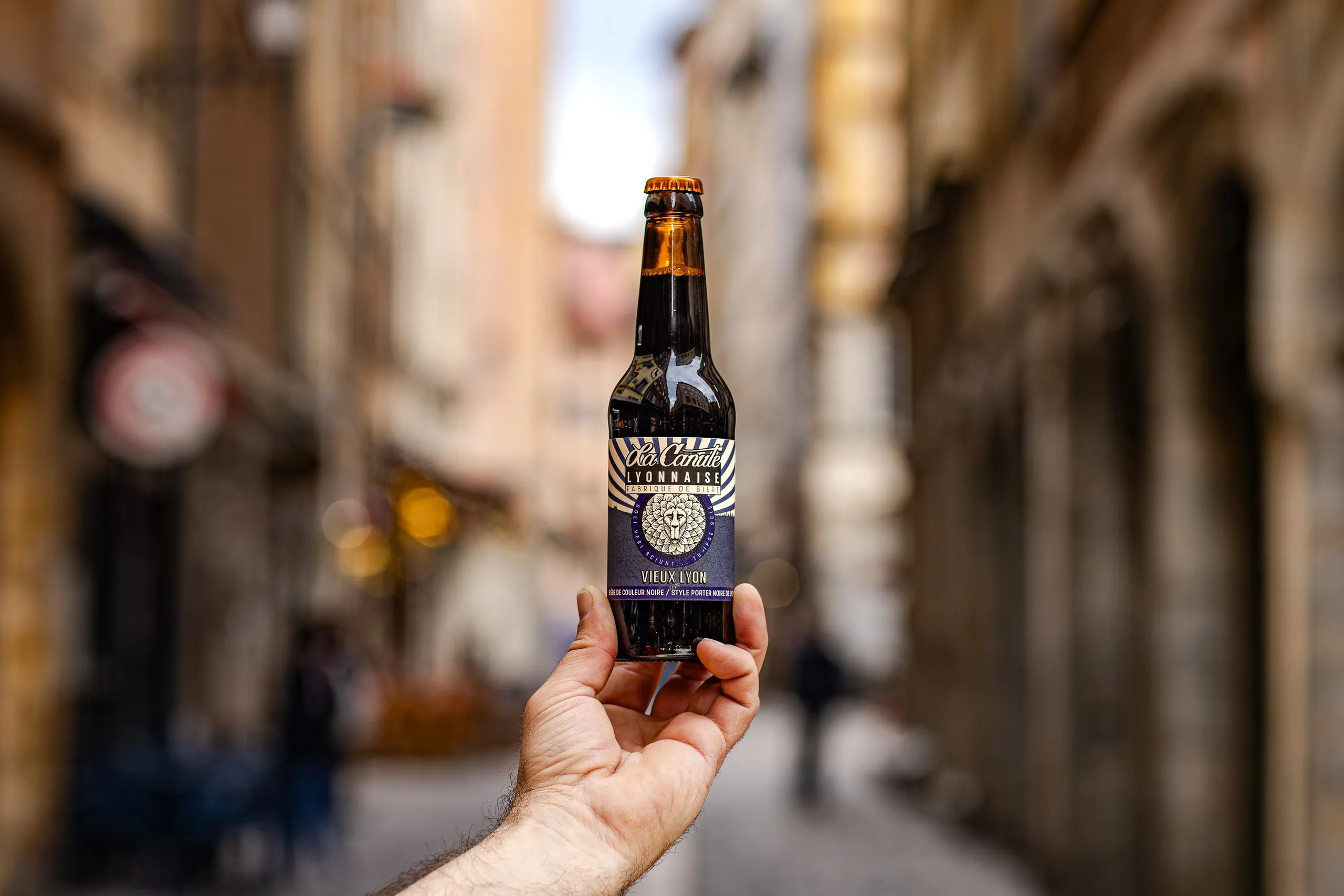 Hand holding up a dark beer bottle in a narrow street of Old Lyon, La Canute Lyonnaise