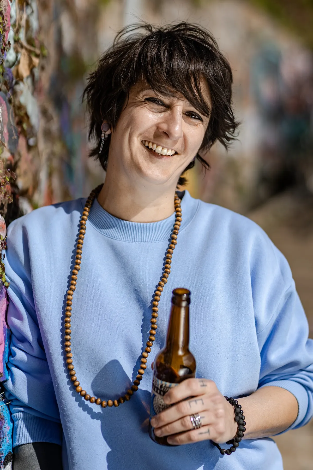 Smiling woman in a blue sweater and a wooden bead necklace holding a bottle, La Canute Lyonnaise team
