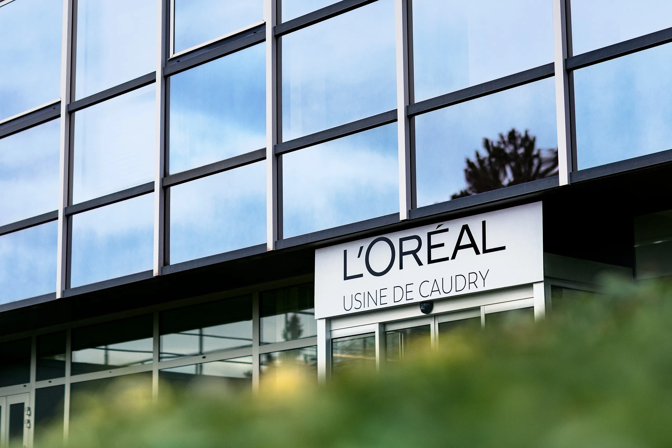 Glass façade of the L'Oréal factory in Caudry behind blurred greenery, Le Monde & L'Oréal