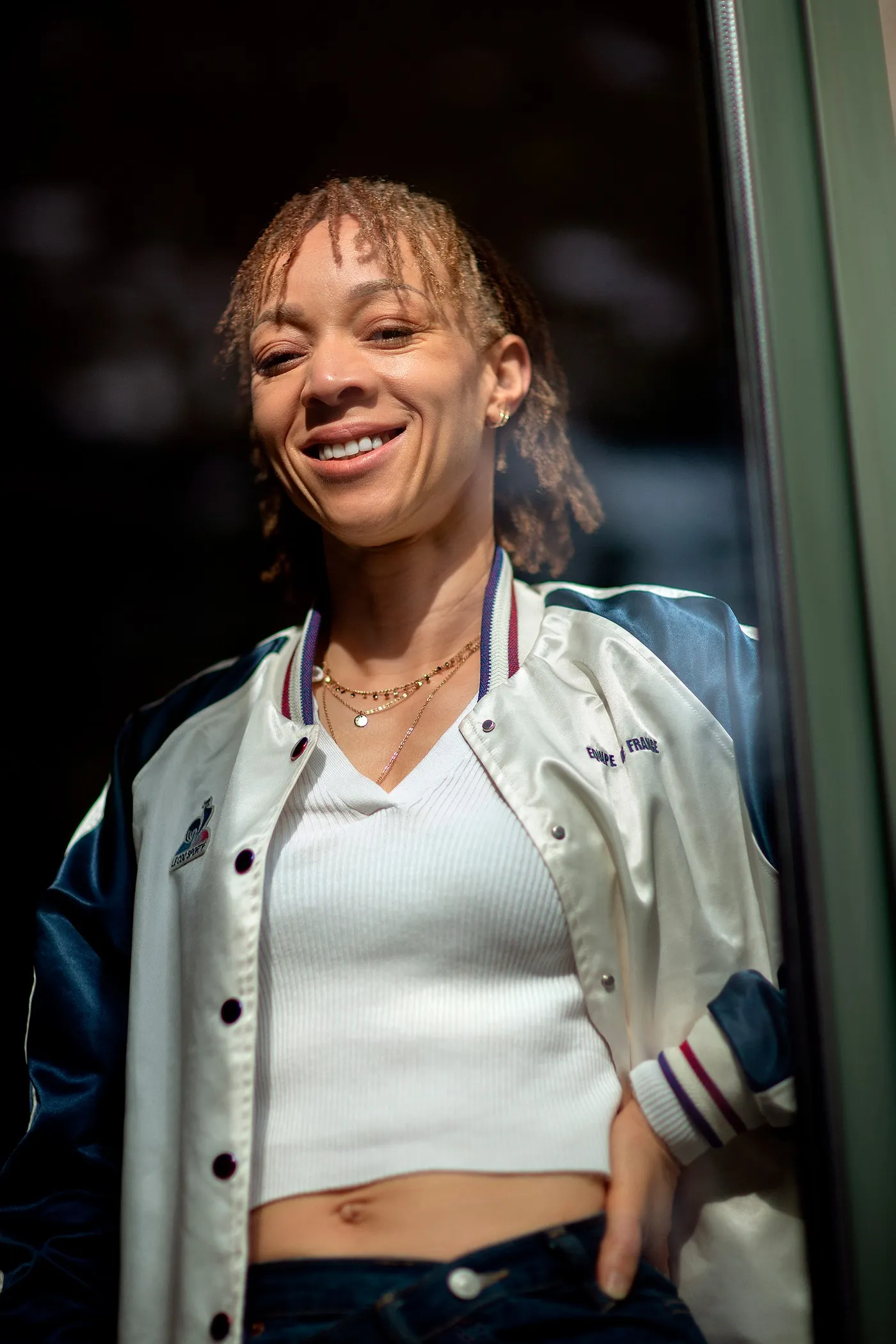 Smiling young woman in a white and navy varsity jacket in a dark doorway, portrait for Le Monde & L'Oréal