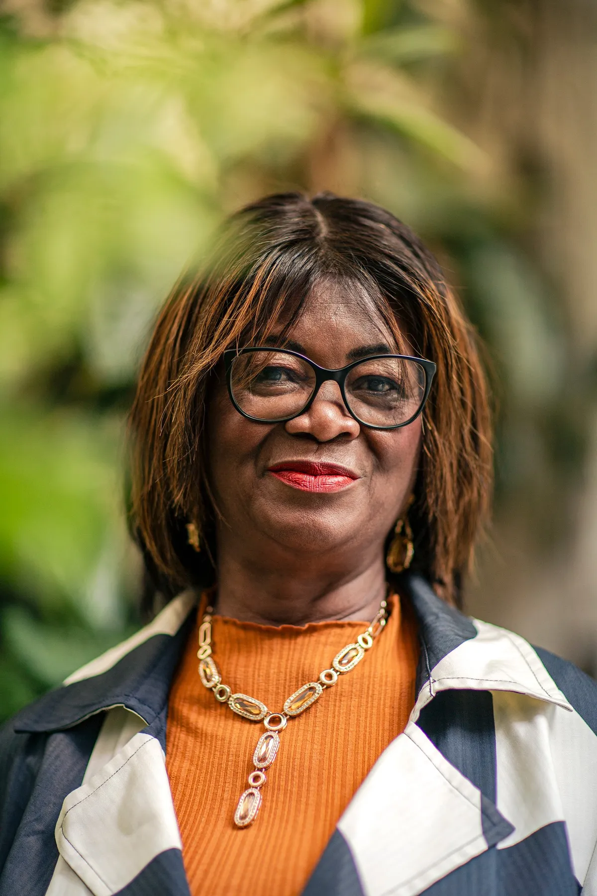 Woman with glasses, an orange top and a gold necklace against green foliage, portrait for Le Monde & L'Oréal