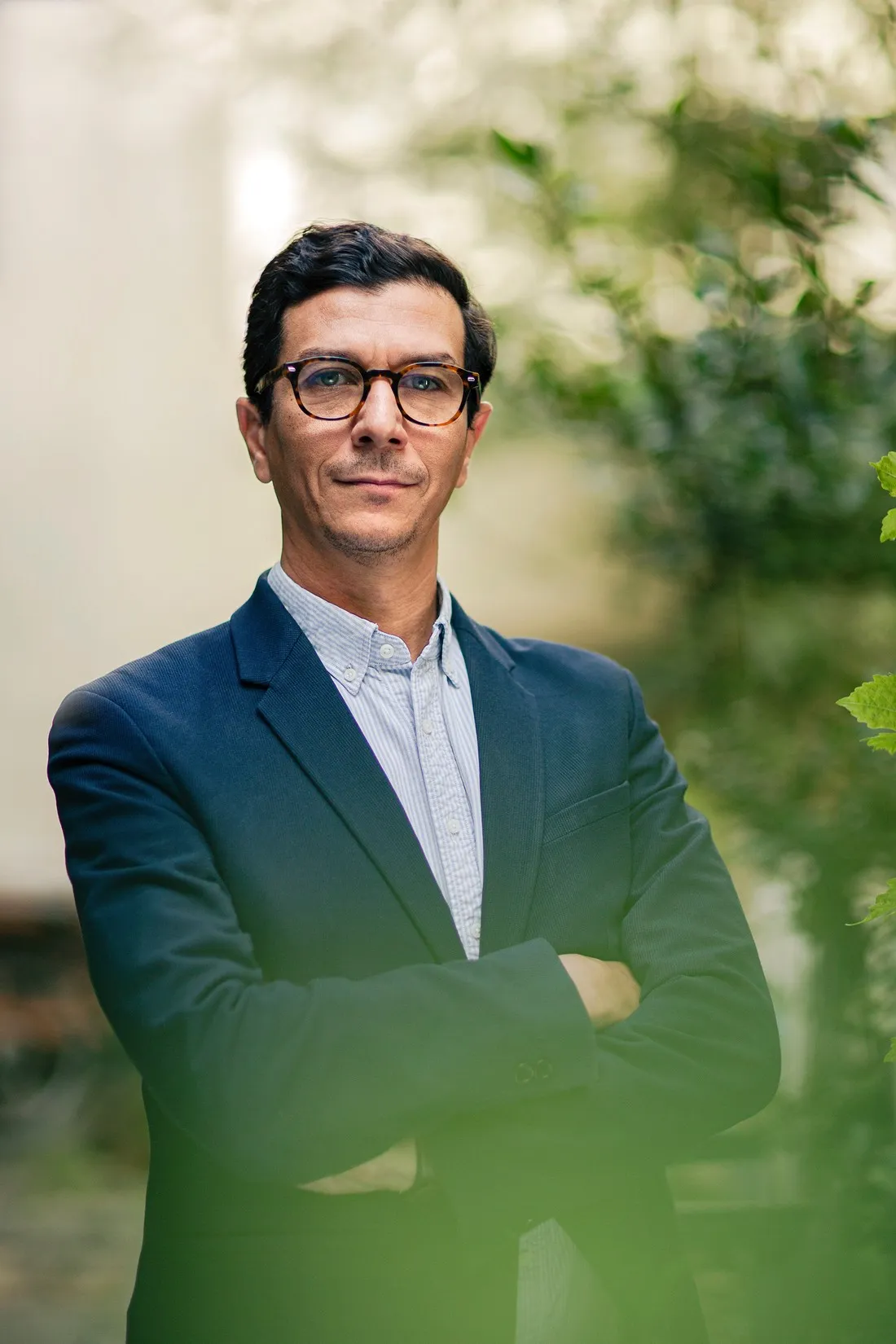 Man with glasses in a navy suit, arms crossed, beside green plants, portrait for Le Monde & L'Oréal