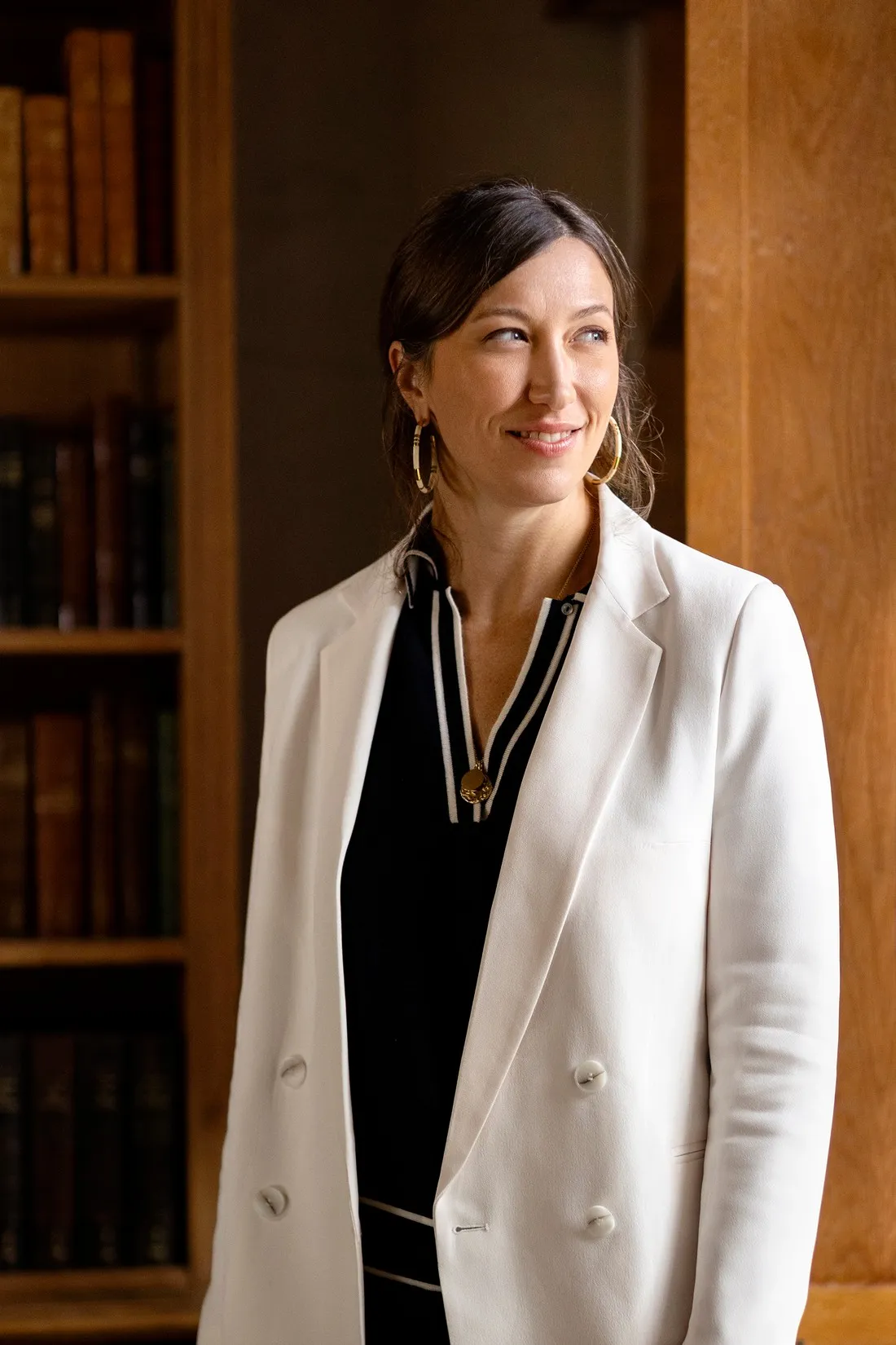 Woman in a white blazer standing by bookshelves in warm light, portrait for Le Monde & L'Oréal