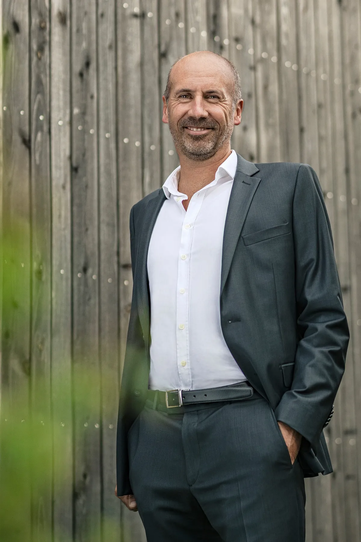 Man in a grey suit and white shirt, hands in pockets, against a wooden fence, portrait for Le Monde & L'Oréal