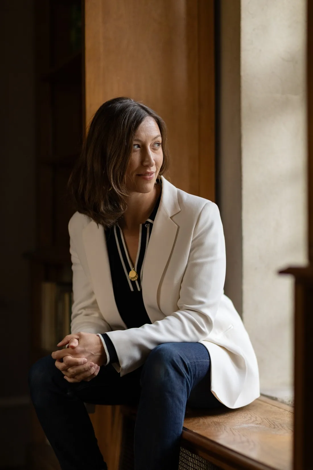 Woman in a white blazer sitting on a window ledge, looking outside, portrait for Le Monde & L'Oréal