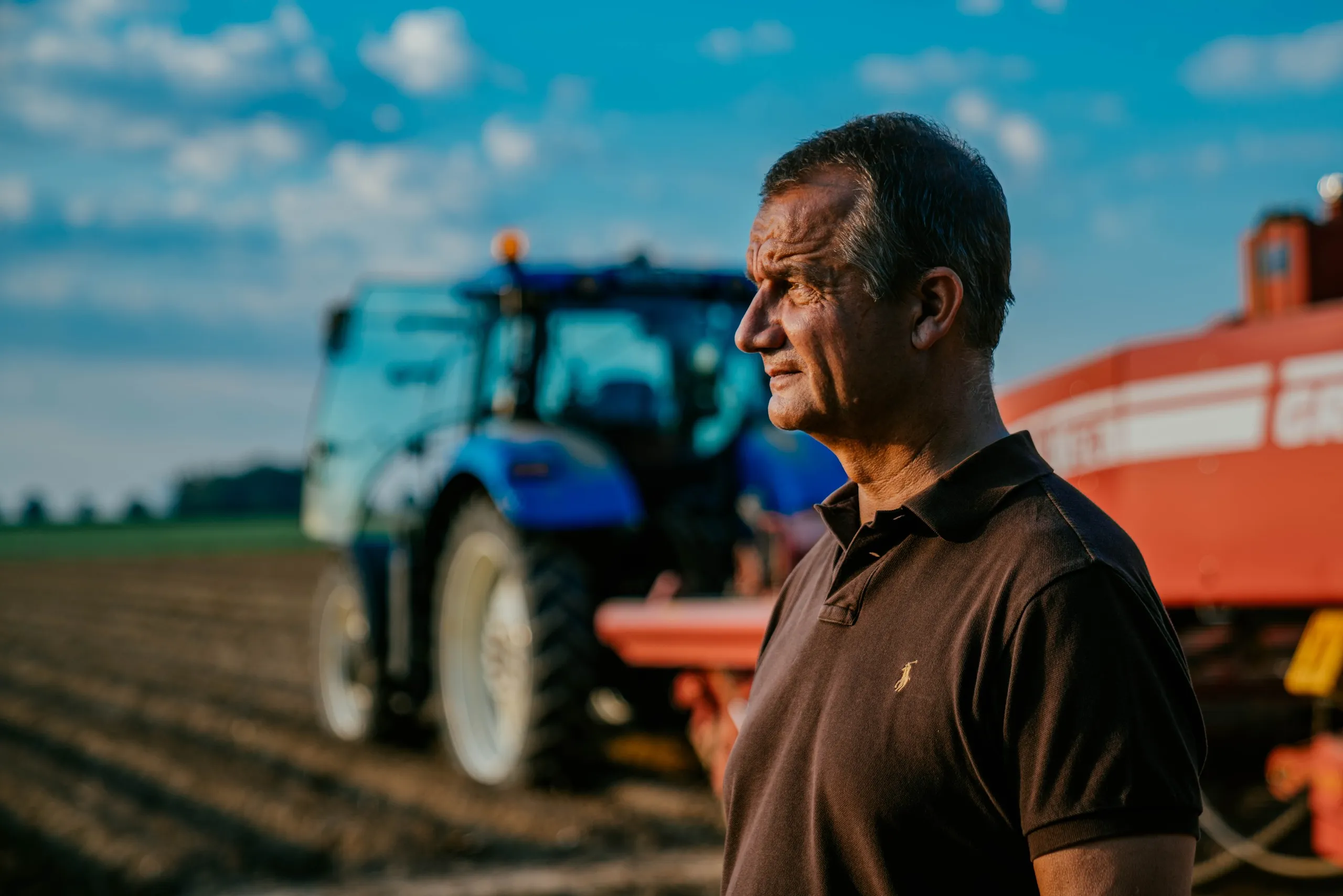 Farmer in a brown polo in front of a blue tractor and a red harvester, McCain farming families