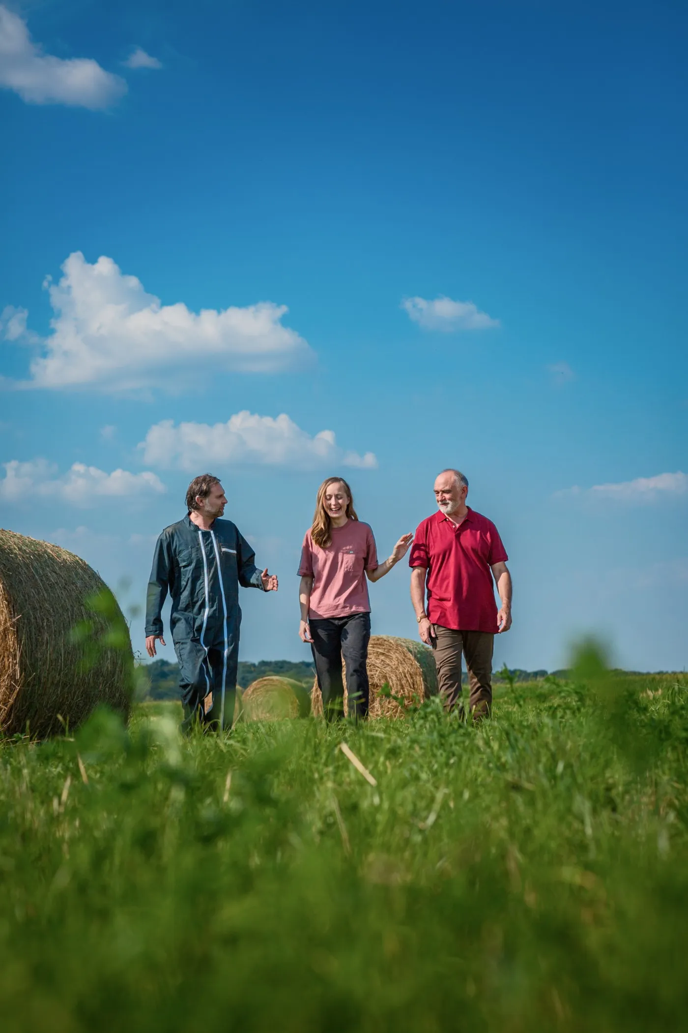 Three people walking through tall grass past hay bales under a blue sky, Mutualité sociale agricole